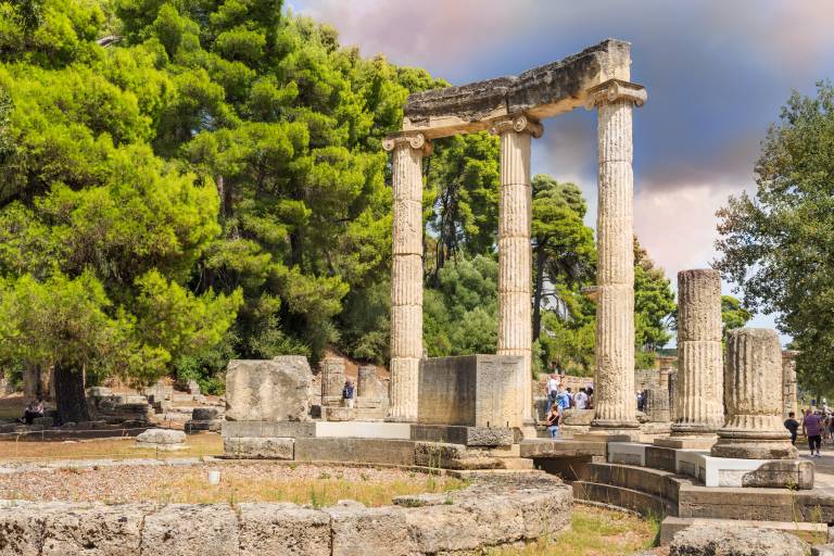 Ancient stone ruins with tall columns stand in the foreground, while lush green trees and a cloudy sky form the background.