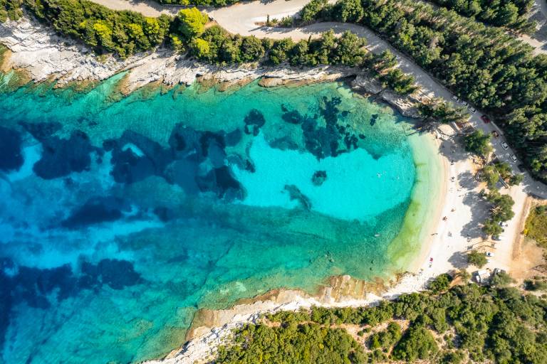 A crescent-shaped sandy beach with turquoise waters in the foreground is surrounded by lush green trees and a winding road in the background.