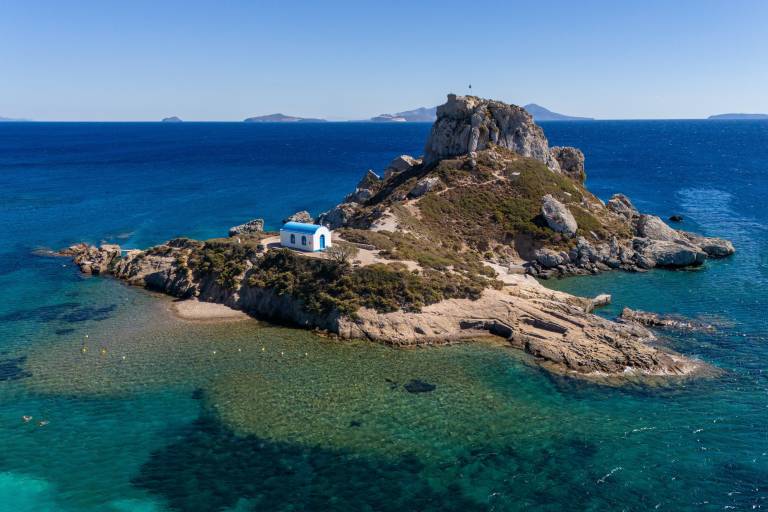 A small white church with a blue roof sits on a rocky island in the foreground, surrounded by clear turquoise water, while distant islands dot the deep blue sea in the background.