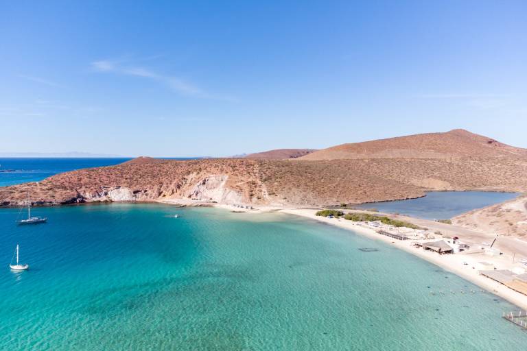 Turquoise waters with sailboats in the foreground lead to a sandy beach and arid hills under a clear blue sky in the background.