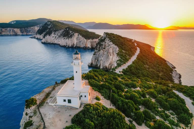 A white lighthouse stands on a rocky, green peninsula jutting into the blue sea, with a golden sunset illuminating the distant islands and horizon.