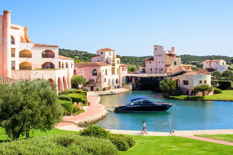 A lone figure strolls along a manicured lawn in the foreground, with a sleek black boat docked in a tranquil canal surrounded by pastel-colored buildings and lush green hills in the background.