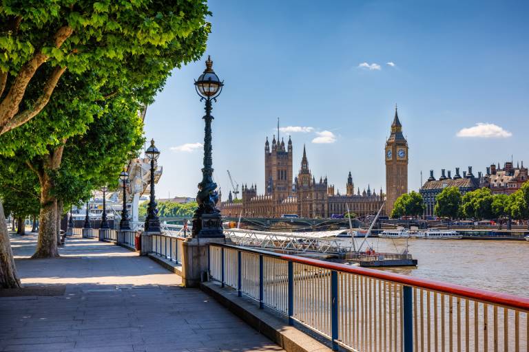 A tree-lined promenade with lampposts in the foreground overlooks the River Thames with the Houses of Parliament and Big Ben in the background.