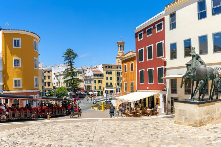 A tourist train is parked in a cobblestone square with colorful buildings and a bronze equestrian statue in the background.