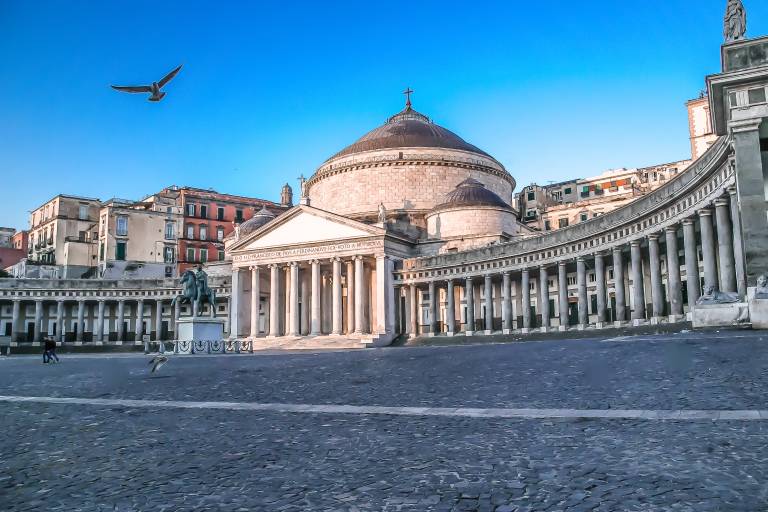 A bird flies over a vast, cobblestone piazza in front of a grand domed building with a colonnade, while apartment buildings rise in the background under a clear blue sky.