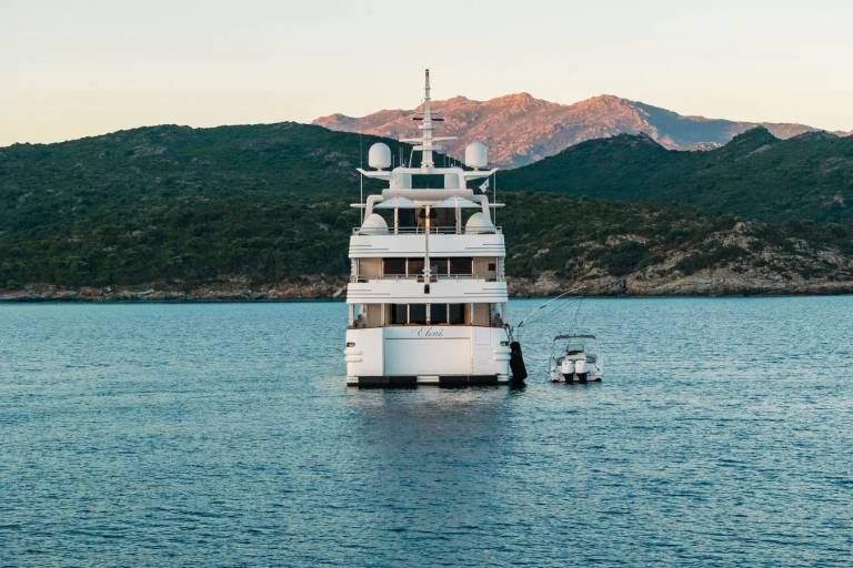 A large white yacht and a smaller boat float on calm blue water in the foreground, with a lush green, mountainous coastline in the background.