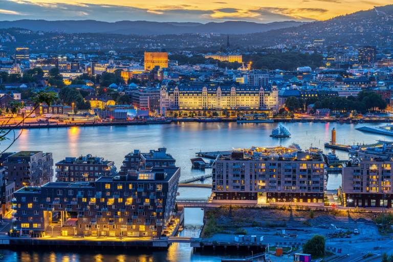 Modern buildings line a waterfront in the foreground, with a sprawling city and hills visible in the background under a twilight sky.