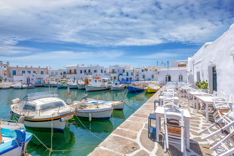 Boats are docked in a harbor with white buildings and outdoor seating in the foreground and background.