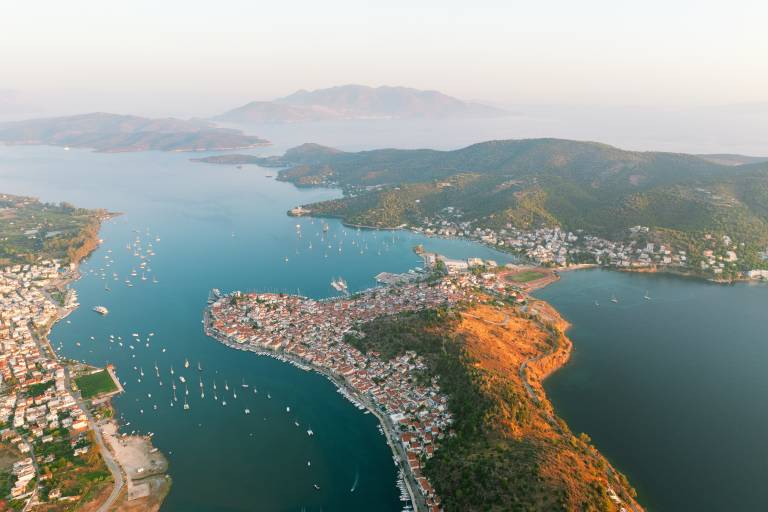 A coastal town with red-tiled roofs and a harbor filled with sailboats is nestled between a calm blue bay and a sun-drenched, tree-covered peninsula, with hazy mountains in the distance.