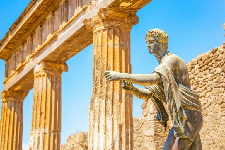 A bronze statue stands in the foreground before ancient Roman columns and ruins under a bright blue sky.