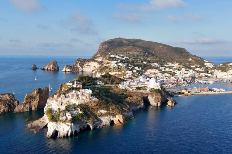 White buildings cling to rocky cliffs in the foreground, while a larger, verdant island with a town and harbor stretches into the blue ocean under a clear sky in the background.