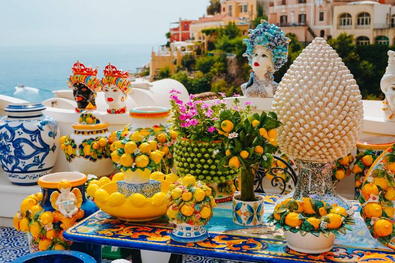 Colorful ceramic vases and lemon-themed decor are displayed on a table in the foreground, with a picturesque Italian coastal town cascading down a hillside in the background.