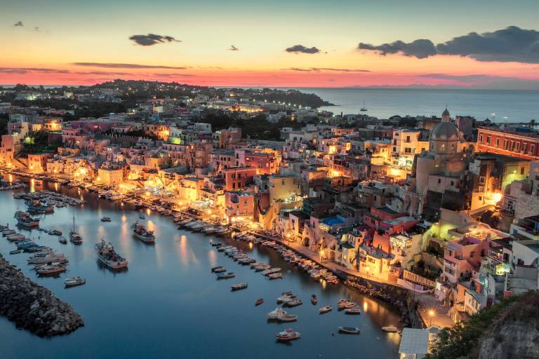 Boats fill a harbor in the foreground, while a colorful, illuminated town stretches towards the sea under a twilight sky in the background.