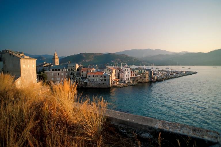 Golden grasses in the foreground frame a picturesque coastal village with a harbor and distant mountains under a clear sky.