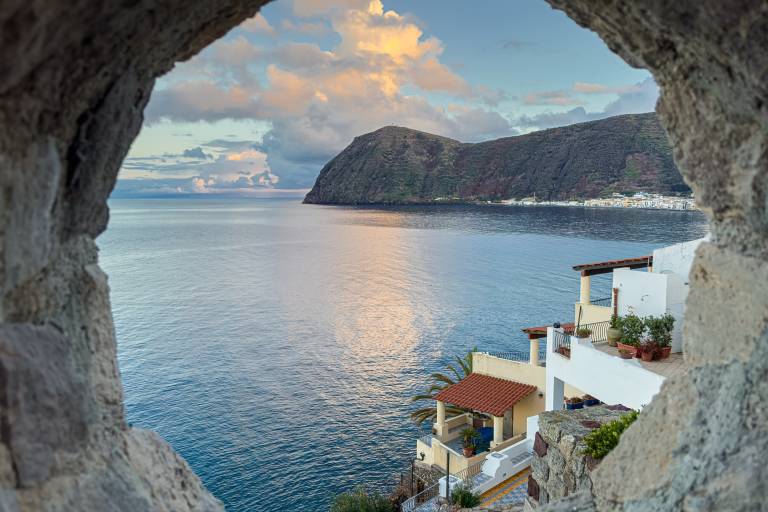 Rough stone frames a tranquil sea with a distant island and white buildings nestled on a cliffside.