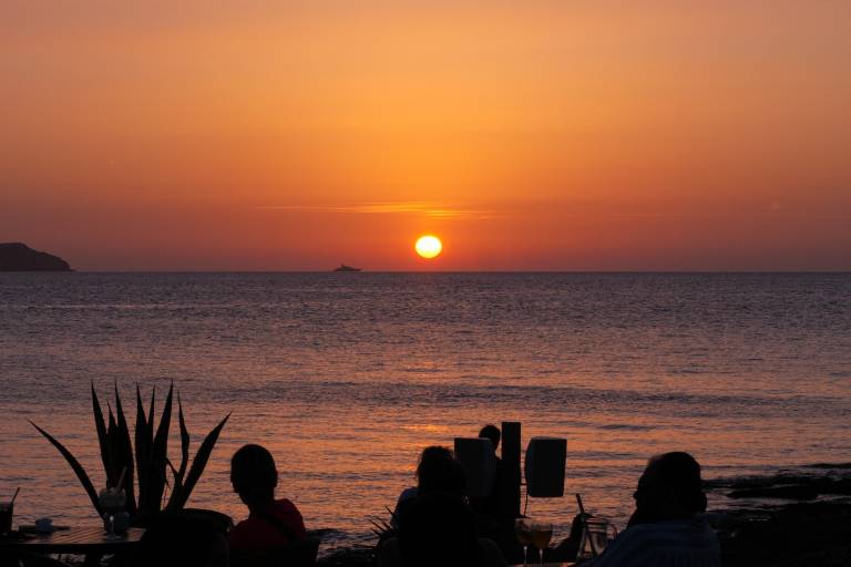 Silhouetted figures and a spiky plant sit at tables in the foreground, while a vibrant orange sunset over the ocean dominates the background.