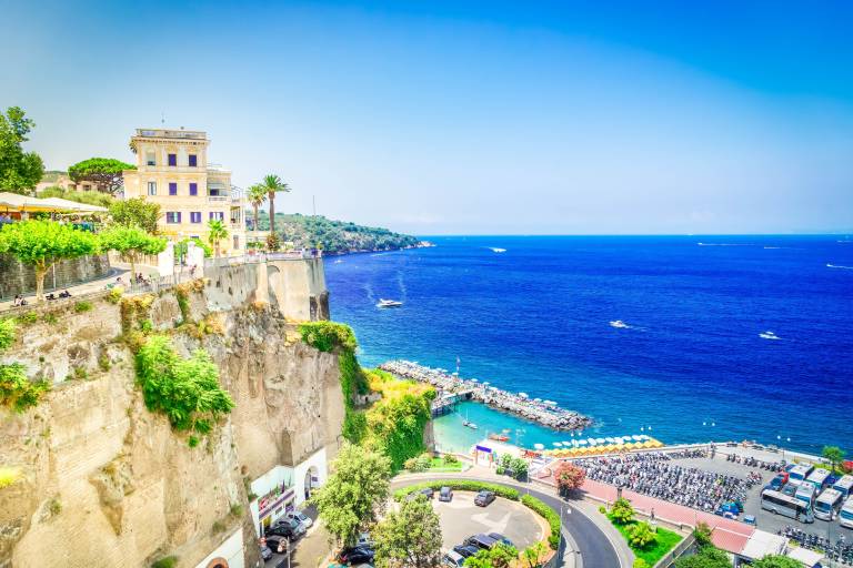 A cliffside town with lush greenery and buildings overlooks a vibrant blue sea dotted with boats and a small harbor.