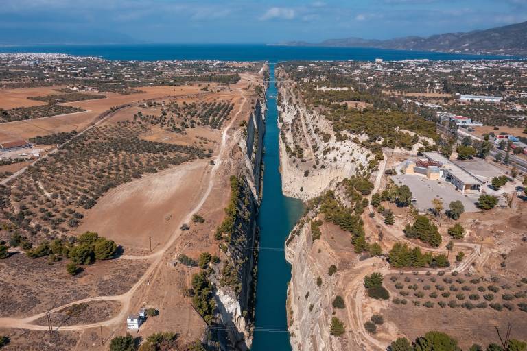 A narrow, turquoise canal cuts through arid, olive-tree-dotted land in the foreground, leading to a distant, hazy coastline with buildings and the sea in the background.