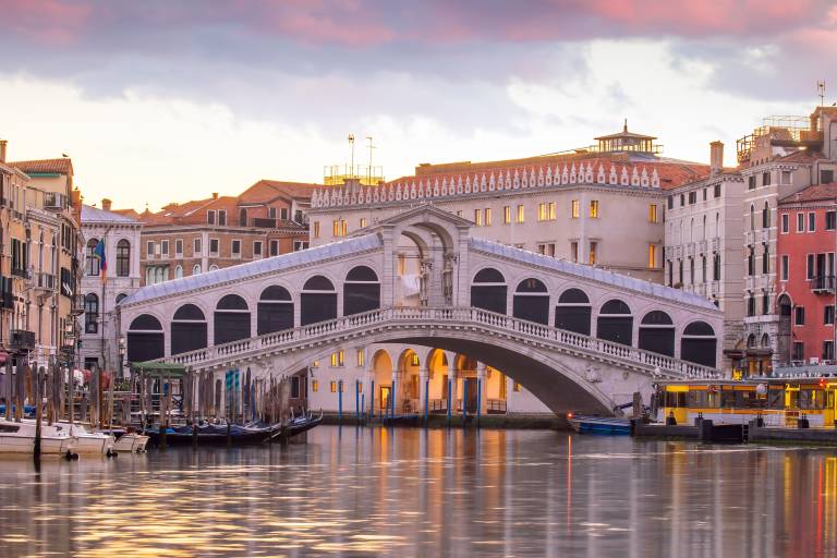 A grand stone bridge arches over a canal in Venice, with colorful buildings and boats lining the waterway under a soft, cloudy sky.