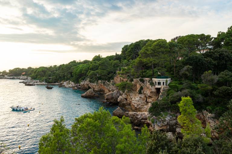 A small boat floats on the calm blue water in the foreground, while a white gazebo sits atop a rocky, tree-covered cliff in the background.