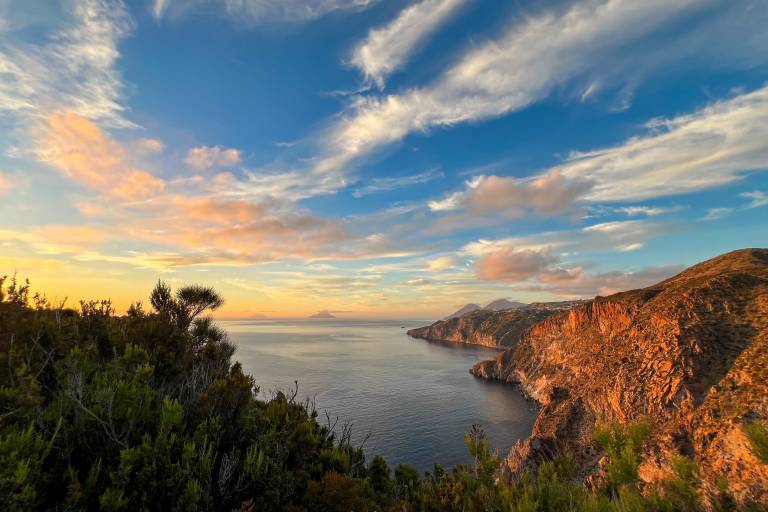 Lush green foliage frames a tranquil bay with rugged, sun-drenched cliffs, all under a vast sky streaked with wispy clouds.
