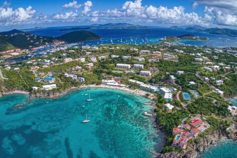 Sailboats dot the turquoise water in the foreground, while a lush, green island dotted with buildings stretches into the background under a bright blue sky.
