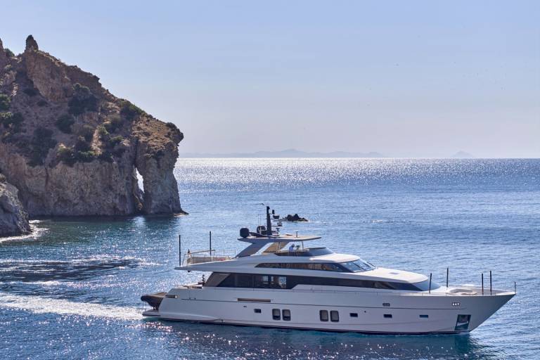 A sleek white yacht cruises through calm blue waters in the foreground, with a dramatic rocky cliff face and the shimmering sea stretching to the horizon in the background.