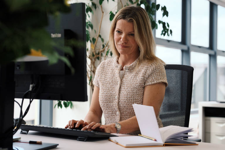 A woman with blonde hair works at a computer in an office with large windows and plants in the background.