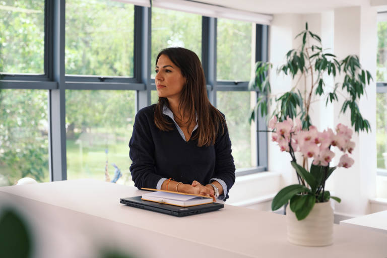 A woman with dark hair looks thoughtfully to the left while sitting behind a white counter with a laptop and notebooks, with a large window and lush greenery in the background.