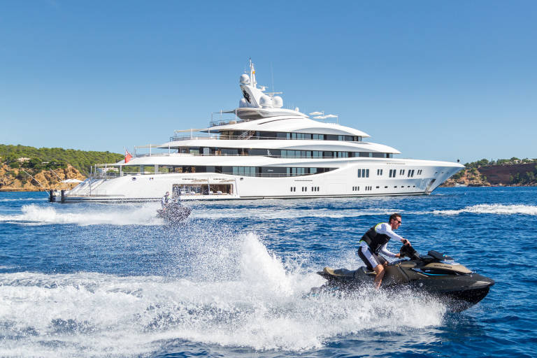 A man rides a jet ski through the blue water, leaving a spray of white foam, with a large white yacht anchored in the background near a tree-covered coastline.