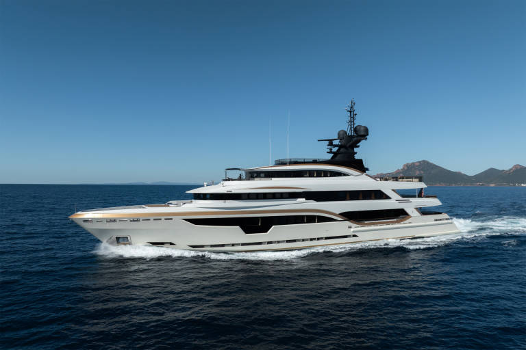 A luxurious white and gold yacht cruises through deep blue water with a distant, hazy coastline under a clear sky.