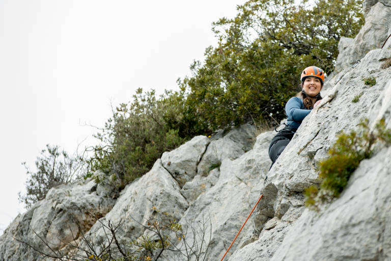 A smiling woman in a blue shirt and orange helmet climbs a rocky cliff face, with green trees and a cloudy sky in the background.