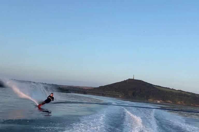 A water skier carves through calm water, leaving a spray of mist, with a tree-covered hill and a distant tower under a clear blue sky in the background.
