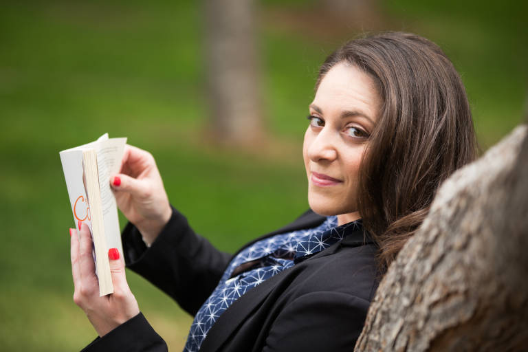 A woman with dark hair and a blue patterned shirt leans against a tree, reading a book, with a blurred green background.