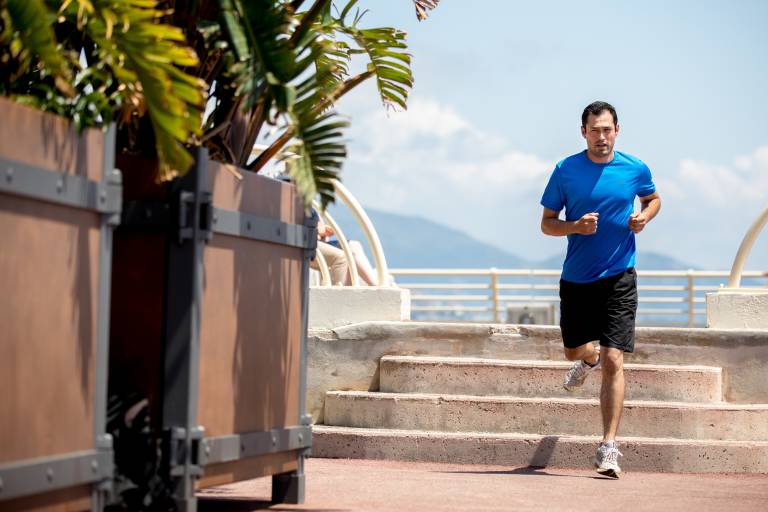A man in a blue shirt and black shorts runs up stairs in the foreground, with lush greenery and a distant coastline in the background.