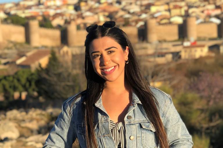 A smiling woman in a denim jacket stands in the foreground with a historic walled city in the background.