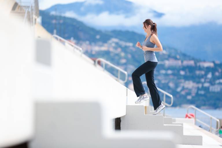 A woman in athletic wear runs up a white staircase in the foreground, with a blurred coastal town and mountains in the background.
