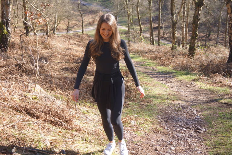 A young woman in a black outfit walks on a dirt path through a wooded area with dry brown foliage in the foreground and bare trees in the background.