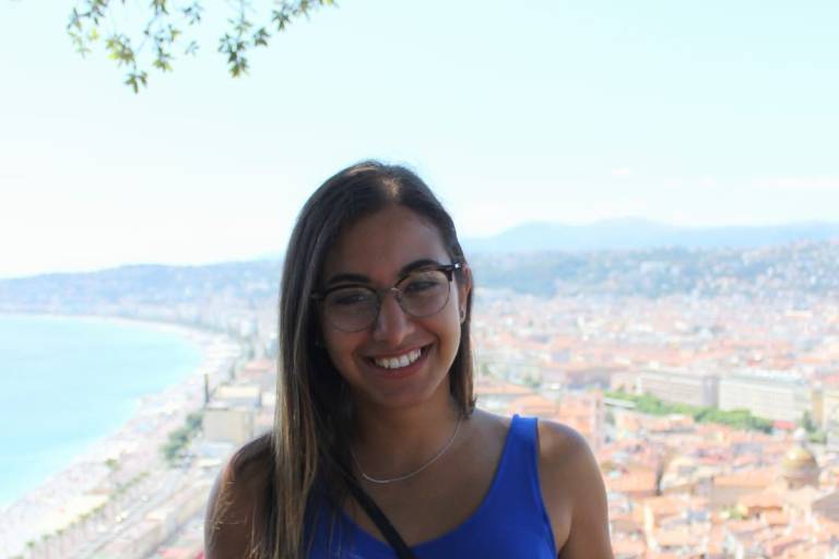 A smiling young woman in a blue dress stands in the foreground with a scenic coastal city and ocean in the background.