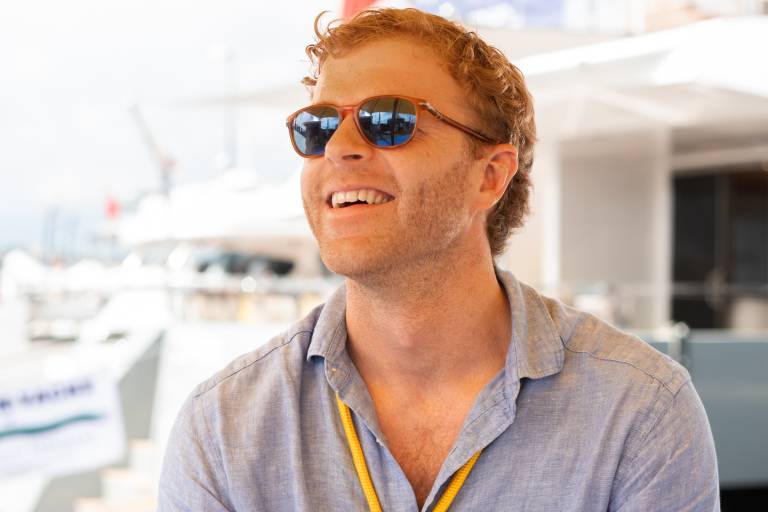 A man with red hair and sunglasses smiles while wearing a blue shirt and a yellow "TEAM" lanyard, with yachts visible in the blurred background under a yellow awning.