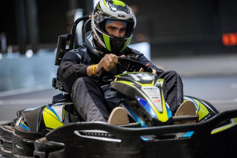 A man in a racing suit and helmet grips the steering wheel of a go-kart on a track, with blurred lights in the background.