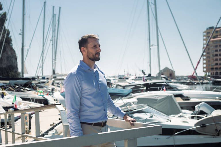 A man in a blue shirt stands on a dock, looking out at a harbor filled with sailboats and yachts under a clear sky.