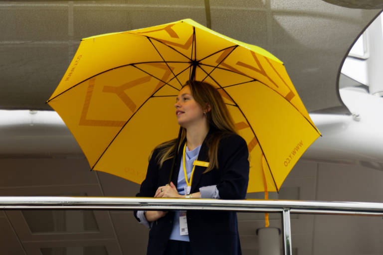 A woman in a dark jacket stands under a bright yellow umbrella with a logo, leaning on a railing with a blurred modern interior in the background.