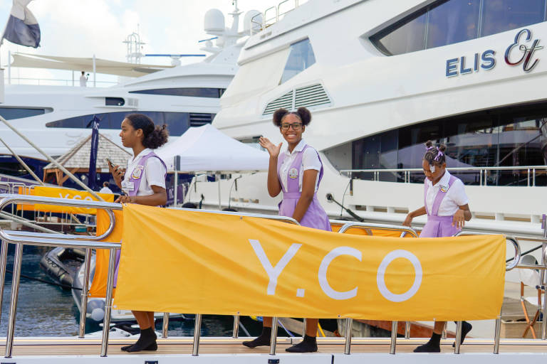 Three young girls in school uniforms stand behind a yellow banner with "Y.CO" printed on it, with large yachts docked in the background.