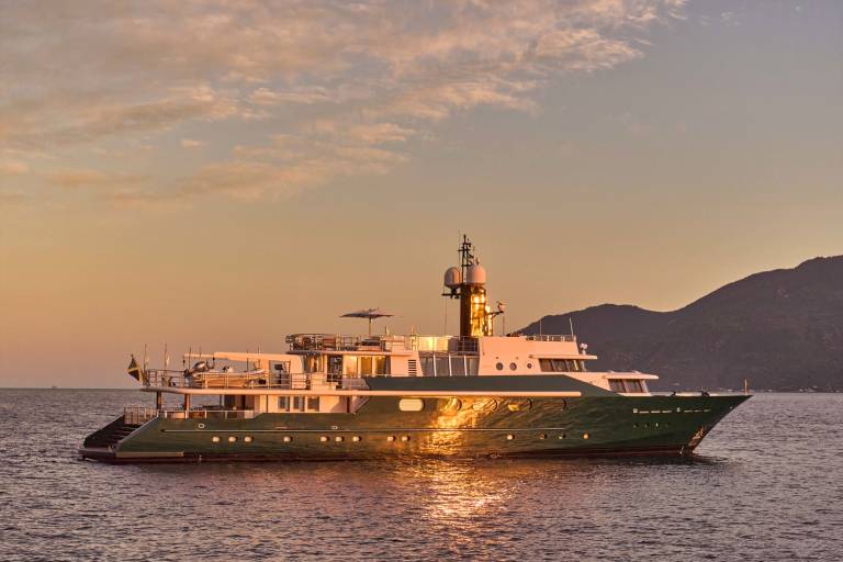 A large green yacht floats on calm water in the foreground, with a hazy, sun-drenched mountain range in the background.