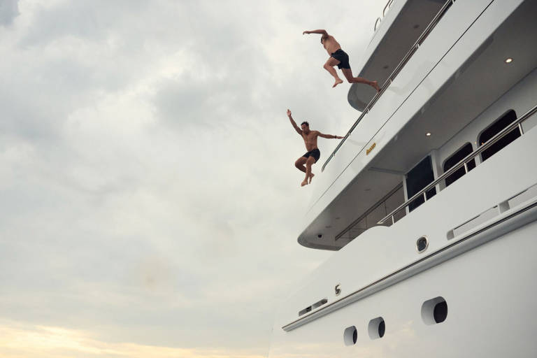Two men leap from the deck of a large white yacht into the cloudy sky.