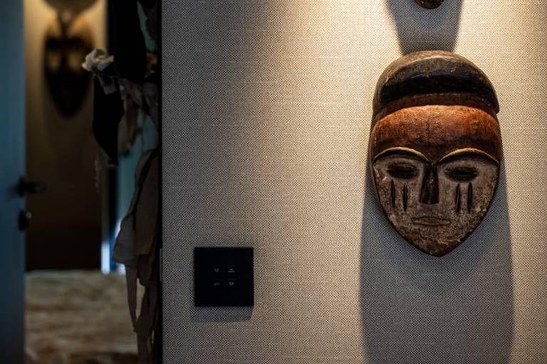 A carved wooden mask hangs on a textured wall in the foreground, with a doorway and blurred interior visible in the background.
