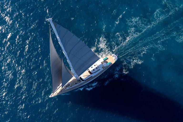 A sleek sailboat with grey sails glides through the deep blue ocean, leaving a white wake behind it.