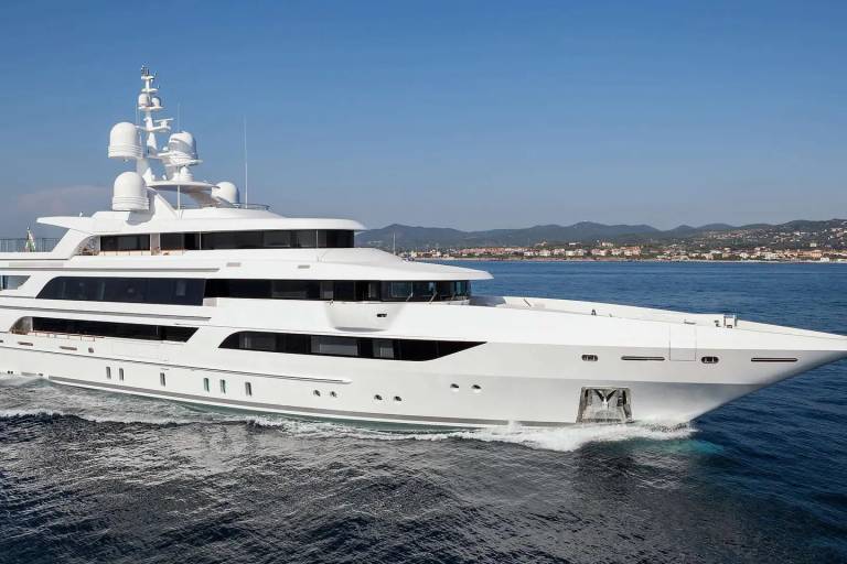 A large, white luxury yacht cruises through deep blue water with a distant coastline and mountains under a clear sky.