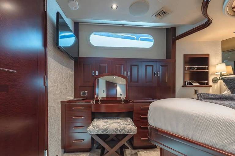 A dark wood door stands in the foreground of a luxury yacht bedroom featuring a vanity with a mirror and a white bed in the background.
